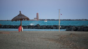 Une plage avec un parasol en chaume, un phare et un mât de drapeau en arrière-plan.