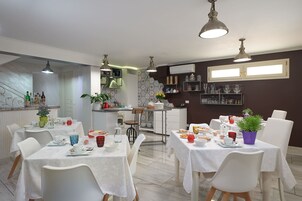 Un comedor con mesas y sillas blancas, una cocina al fondo y plantas decorativas.