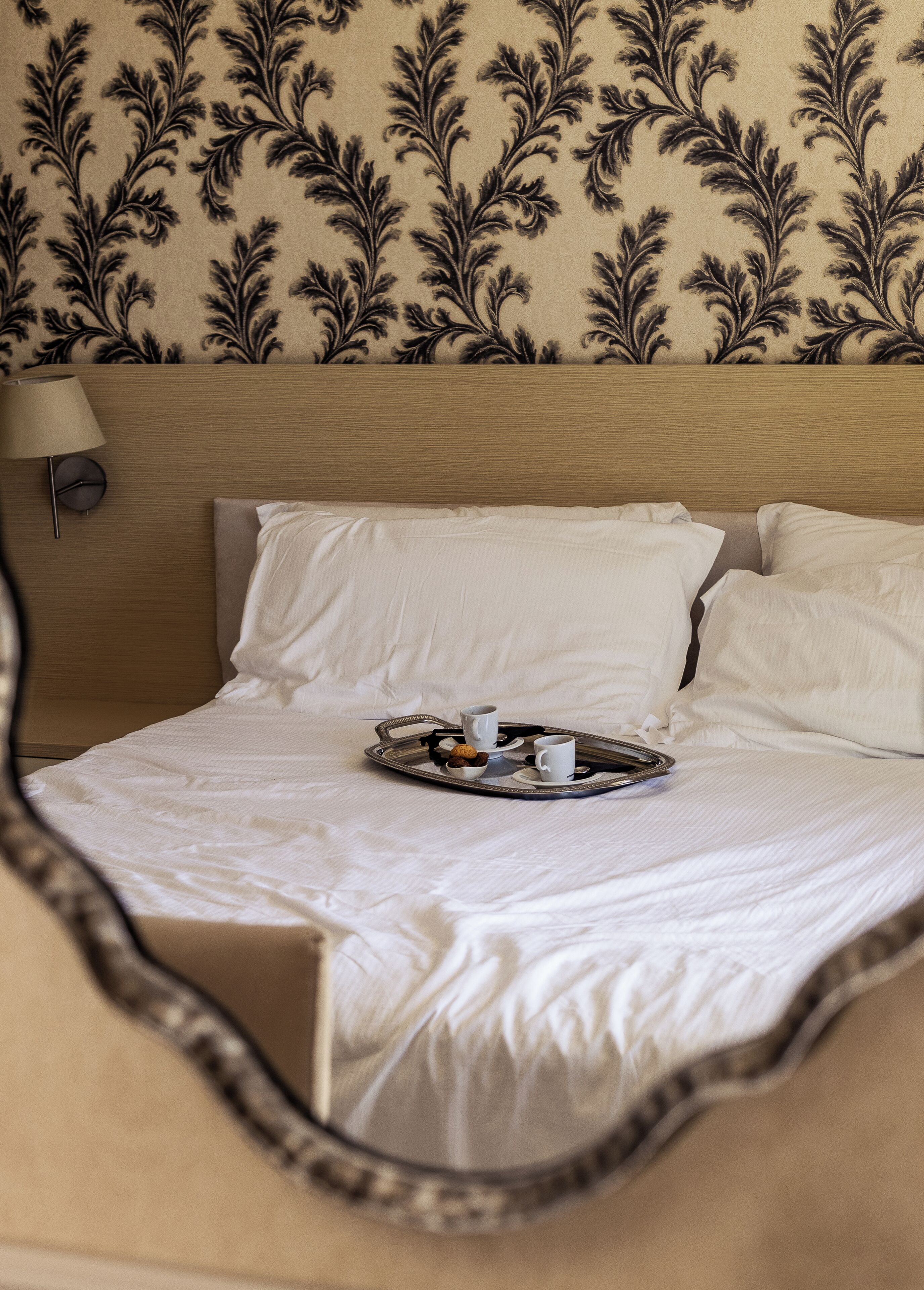 A neatly made bed with white linens, a tray with two cups and a bowl of nuts, and a wall with a floral pattern.