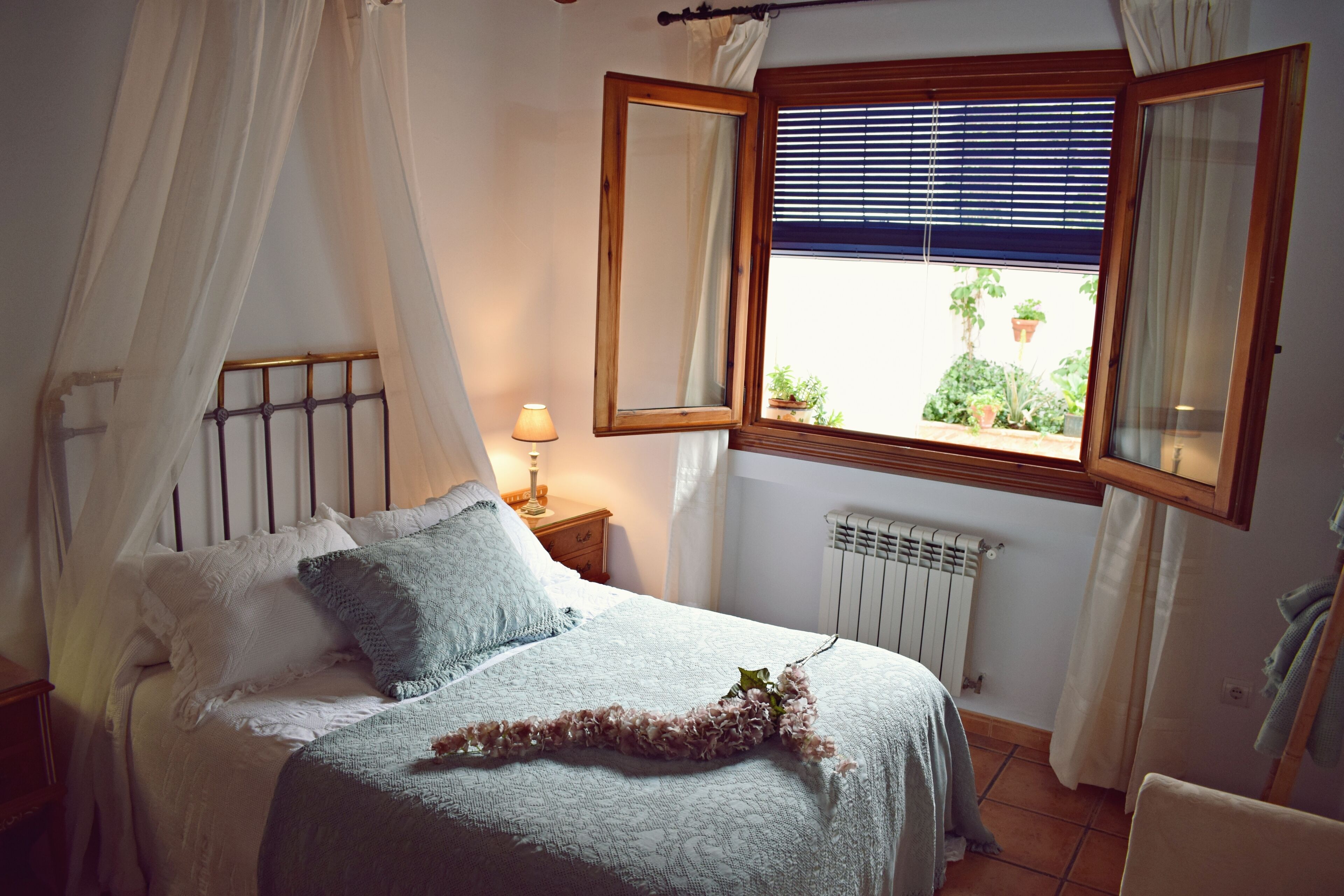 Un dormitorio con cama, mesita de noche, ventana con persianas y radiador.