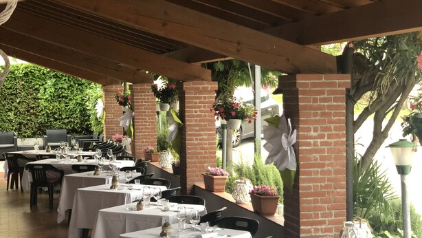 Un espacio de comedor al aire libre con mesas dispuestas para dos, cubierto por una pérgola de madera.