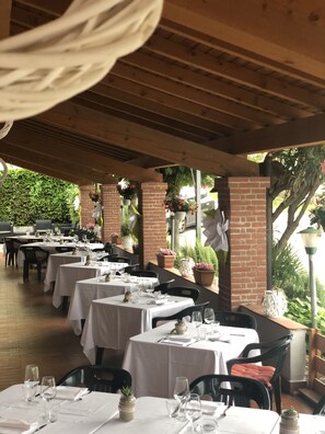 Área de refeições ao ar livre com mesas preparadas para dois, coberta por uma pérgula de madeira.