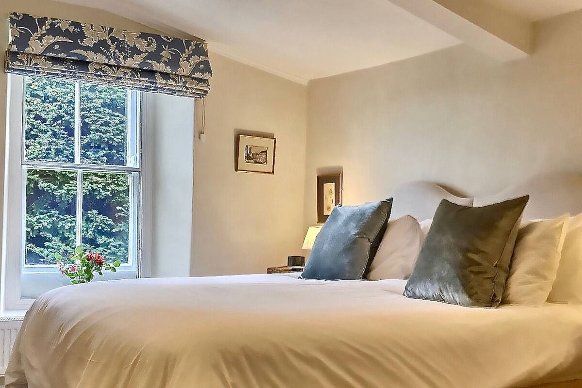 A large bed with a beige bed skirt, a window with a patterned valance, and a framed picture on the wall.