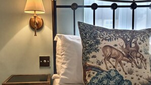 A bedroom with a bed, a nightstand, a lamp, a window with a floral view, and a decorative pillow with a nature scene. - Grove House (Leyburn)