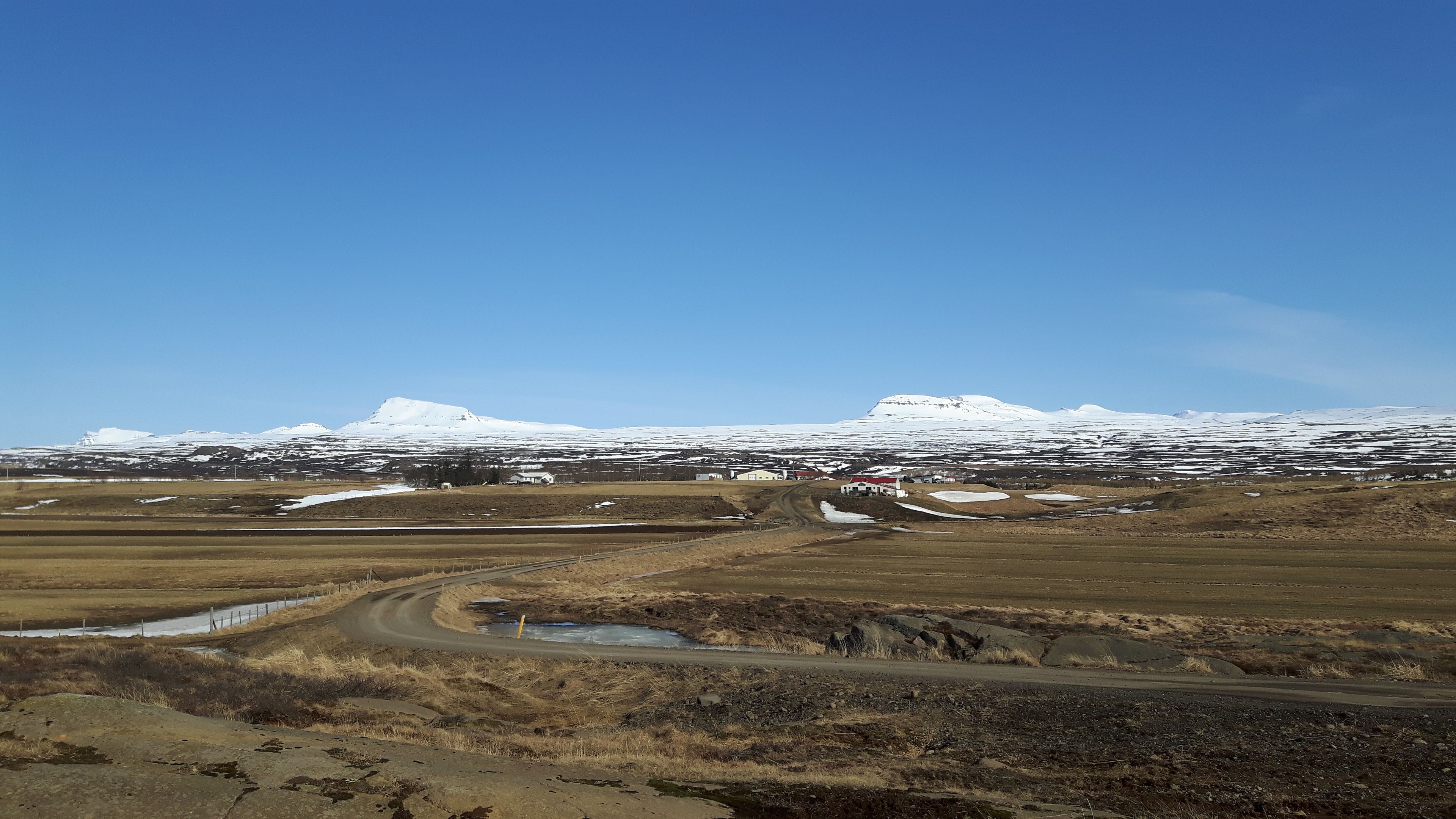 A rural landscape with a dirt road leading to a small settlement. Snow-capped mountains are visible in the background.