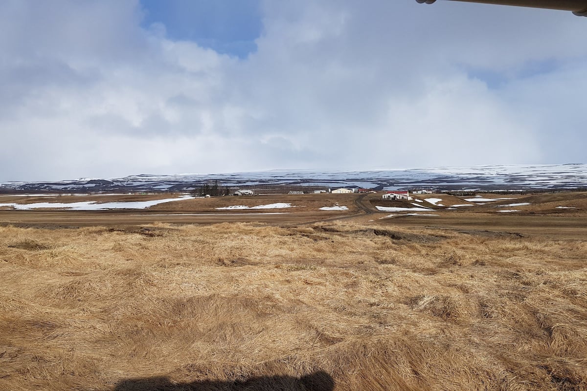 Paisaje rural con cielo despejado, parches de nieve y una edificación a lo lejos.