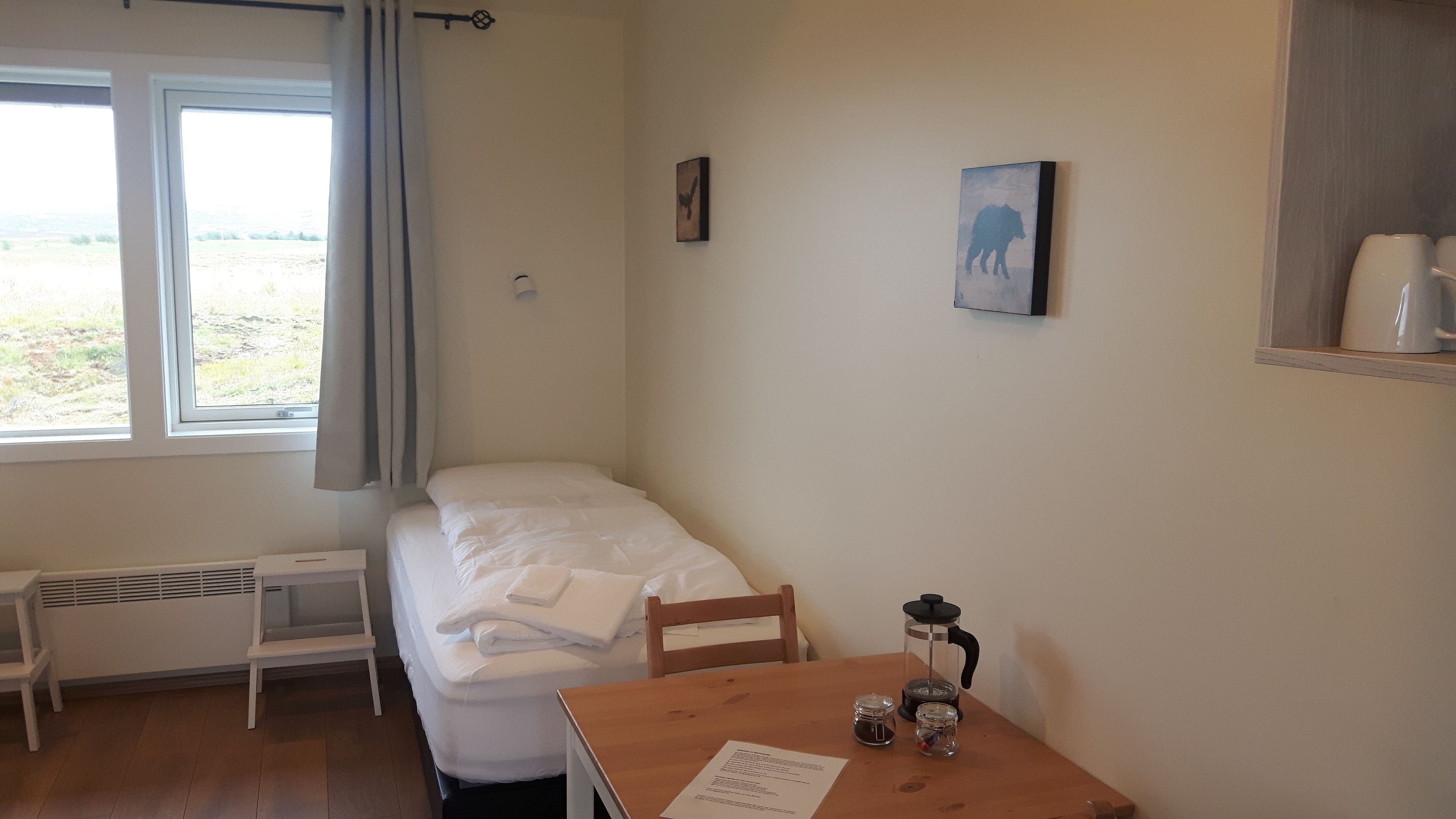 A small, tidy room with a single bed, a wooden table with a coffee maker, and a window with curtains.