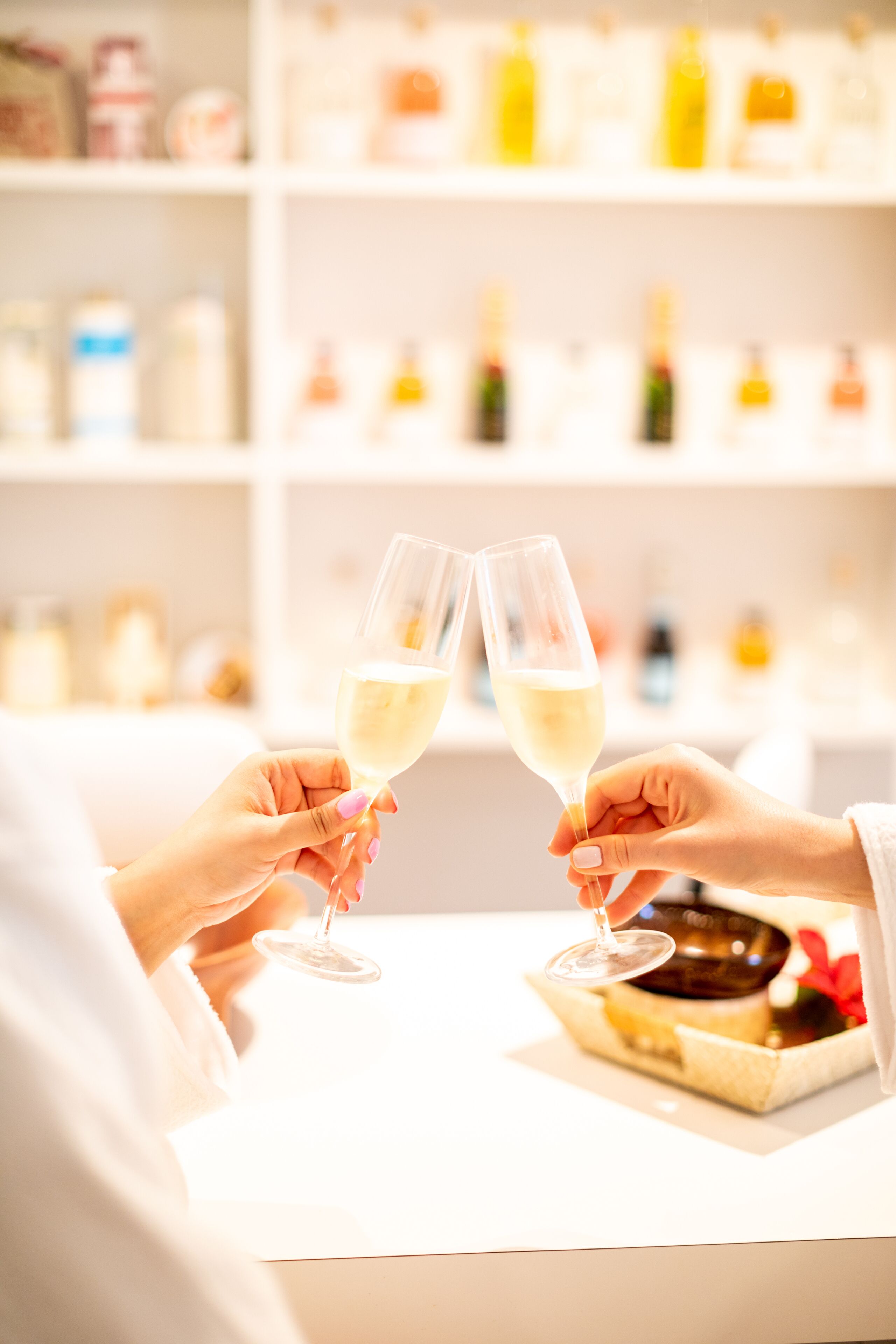 Two hands holding wine glasses, a white table, and a shelf with various bottles in the background.