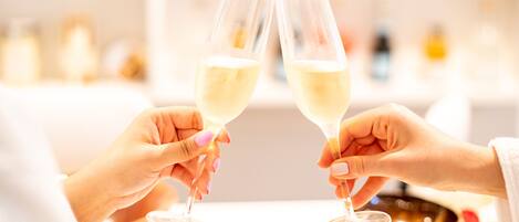 Two hands holding wine glasses, a white table, and a shelf with various bottles in the background.