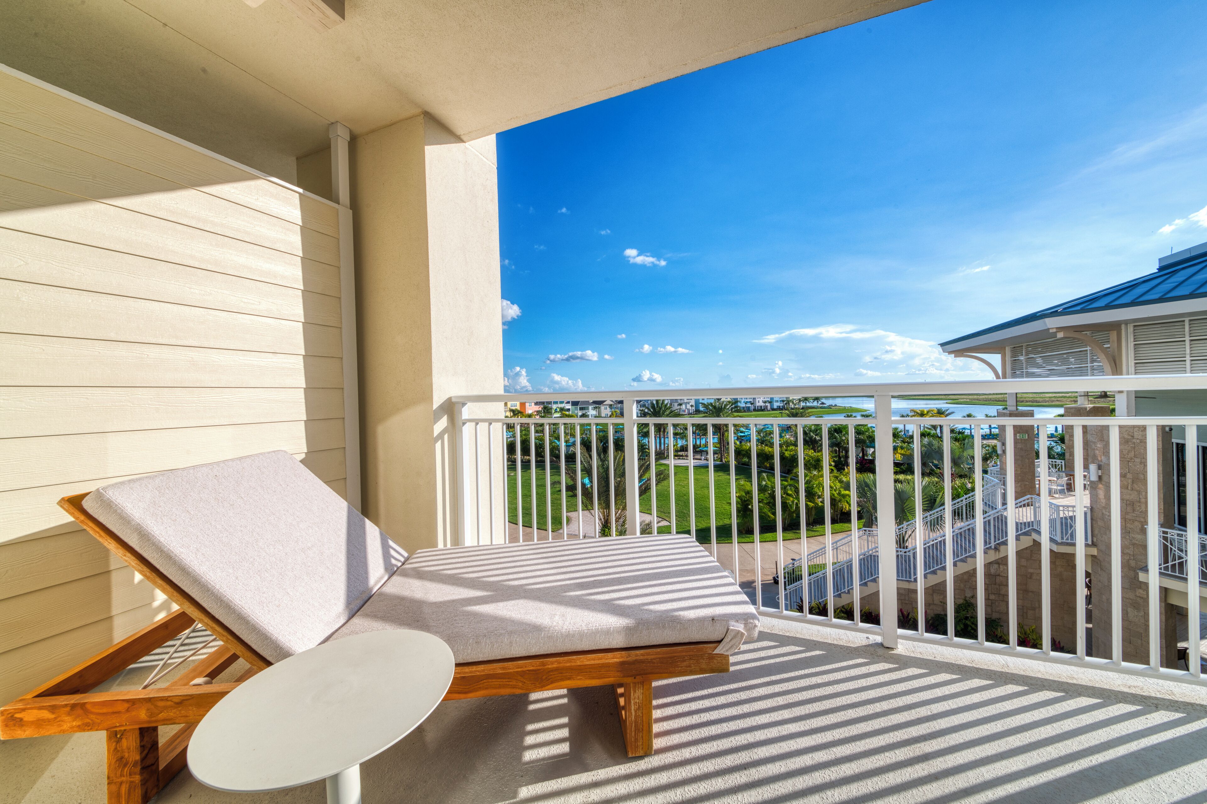 A balcony with a wooden lounge chair and a white table, overlooking a landscaped area with a pool and a building in the background.