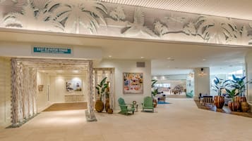 A spacious lobby with a sign for the East Elevator Tower, featuring a tropical mural, seating areas, and potted plants.