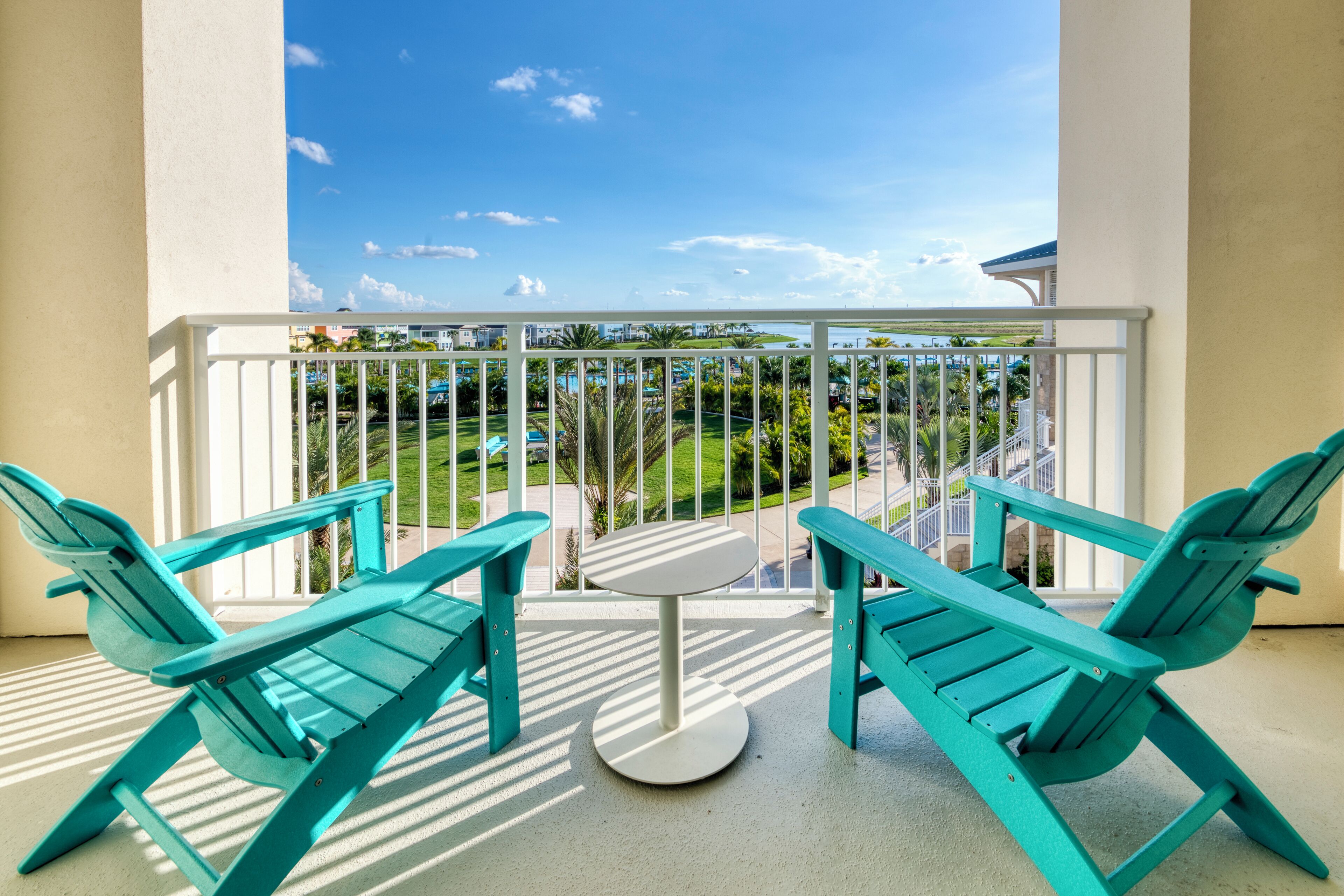 A balcony with turquoise chairs and a small table, overlooking a pool and greenery.