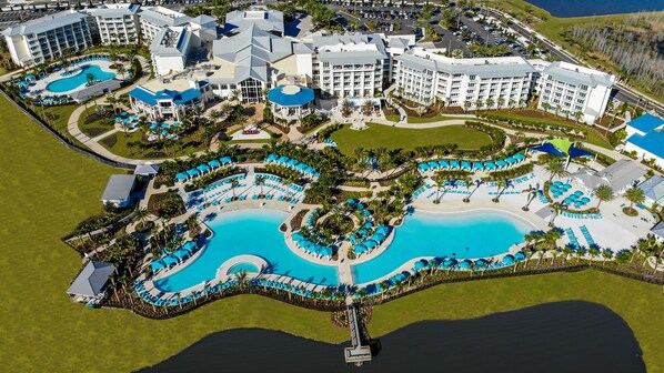 An aerial view of a resort with multiple swimming pools, connected by walkways, surrounded by lush greenery and palm trees.