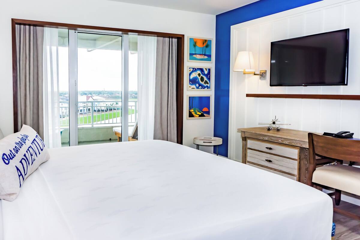 Ein Hotelzimmer mit einem großen Bett, einem Schreibtisch, einem Sessel, einem Fernseher und Blick auf die Stadt durch das Fenster.