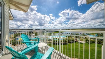A balcony with turquoise Adirondack chairs, a view of a pool and resort area, and a clear sky with clouds.
