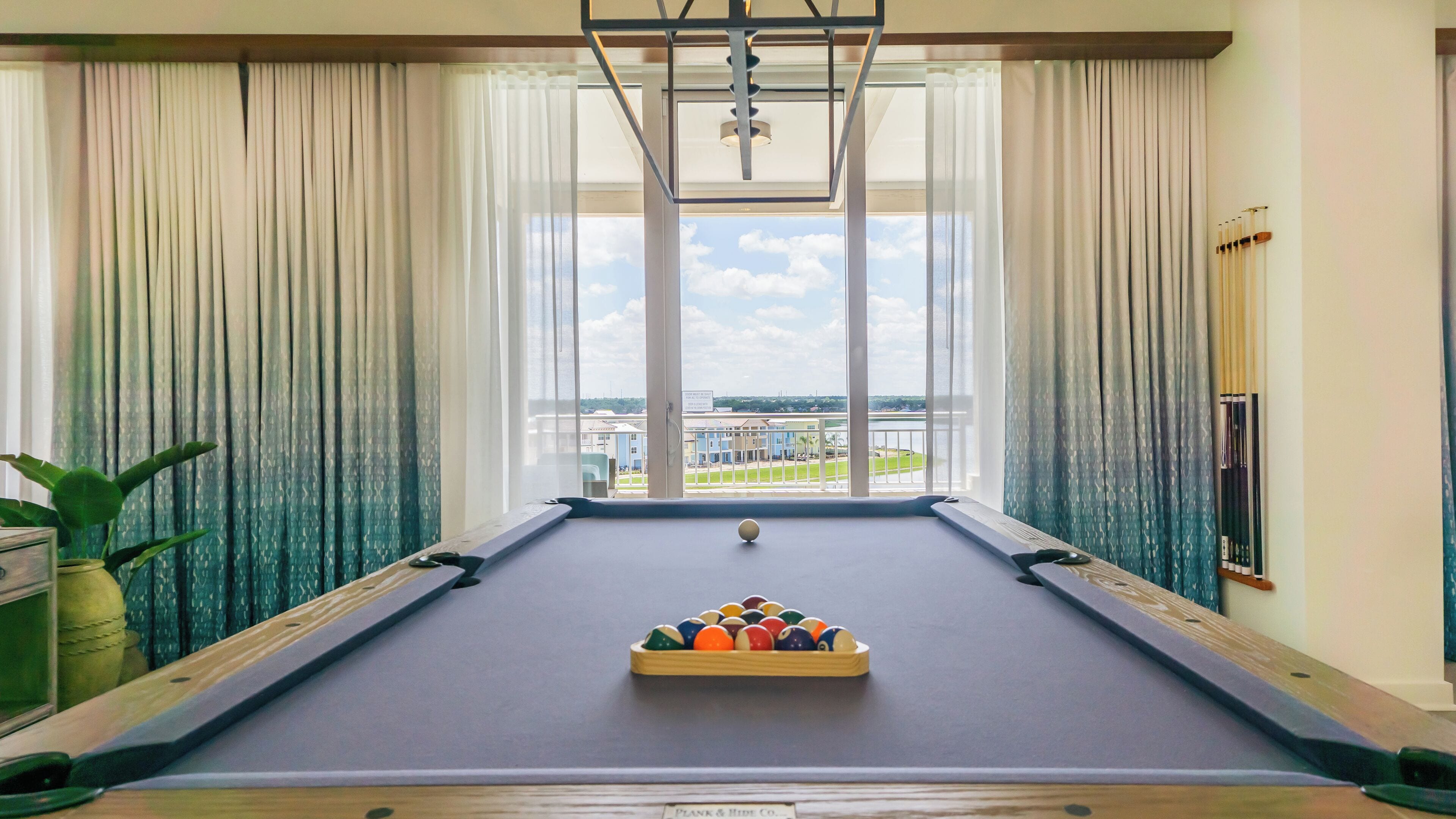 A pool table with a cue ball and a rack of billiard balls, set against a backdrop of a large window with curtains, overlooking a scenic view.