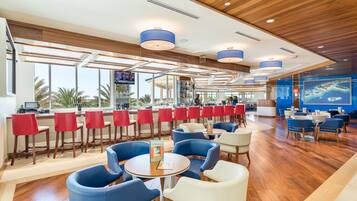 Un restaurante moderno con techos de madera, sillas azules y rojas, y grandes ventanales con vistas a palmeras.