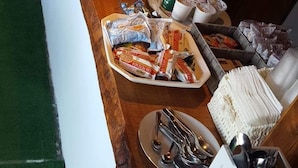 A buffet setup with various food items, including fruits, pastries, and beverages, along with serving utensils and a coffee machine.
