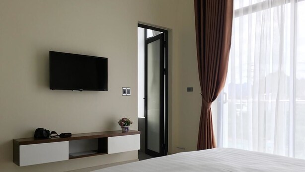 A hotel room with a bed, a flat-screen TV mounted on the wall, a small bedside table with a plant, and a sliding door to the outside. - Ben's Hotel (Thao Nguyen)
