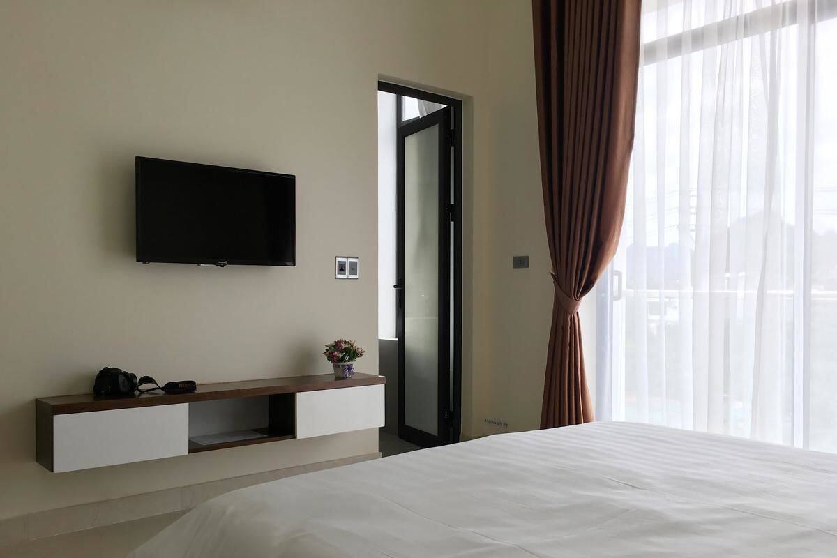 A hotel room with a bed, a flat-screen TV mounted on the wall, a small bedside table with a plant, and a sliding door to the outside.