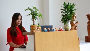 A woman in a red dress standing at a wooden desk with a laptop, surrounded by decorative items including figurines and potted plants. - Ben's Hotel (Thao Nguyen)