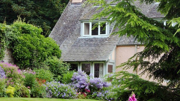 A traditional house with a sloped roof, a chimney, and a garden with various flowers and shrubs. - Finzean Estate Holiday Cottages (Banchory)
