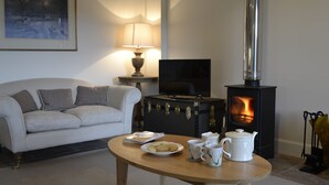 A cozy living room with a sofa, a table with a plate of cookies, a television, and a wood-burning stove. - Finzean Estate Holiday Cottages (Banchory)
