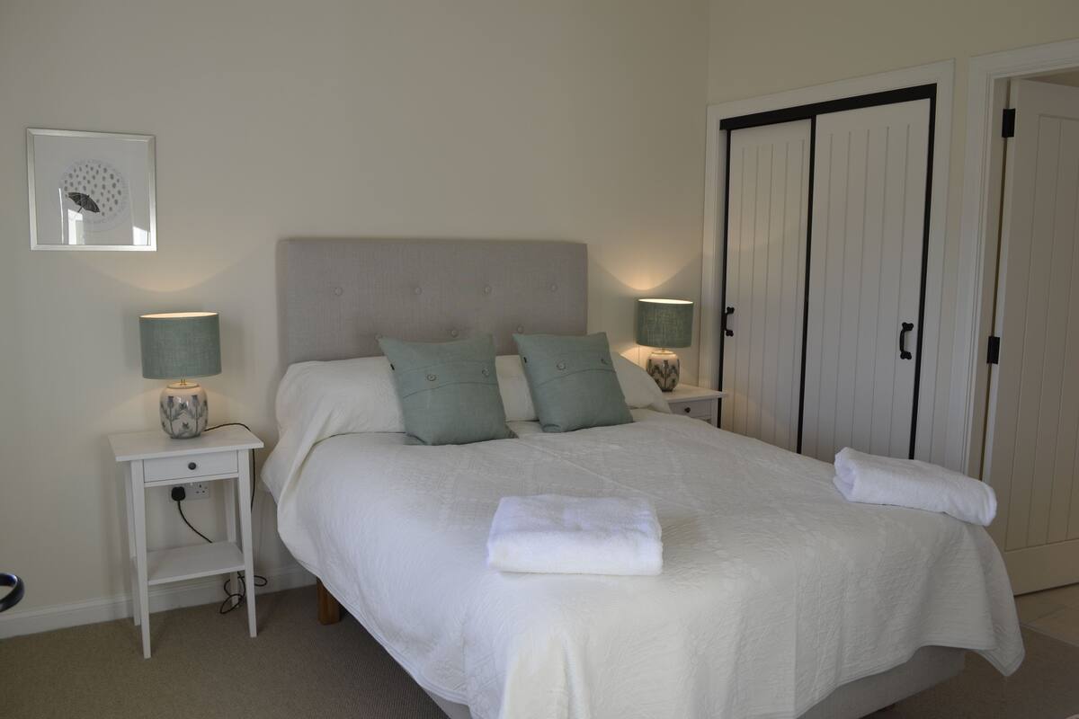 A neatly made bed with two pillows, a bedside table with a lamp, and a wardrobe with a door.