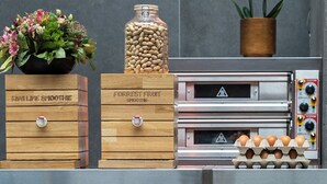 Una zona de cocina con fondo de pared de ladrillo, una lámpara colgante, una caja de madera con la etiqueta 'FORREST FRUIT SMOOTHIES', un tarro de cacahuates, una orquídea en maceta y una pila de platos.