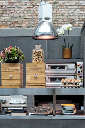 Una zona de cocina con fondo de pared de ladrillo, una lámpara colgante, una caja de madera con la etiqueta 'FORREST FRUIT SMOOTHIES', un tarro de cacahuates, una orquídea en maceta y una pila de platos.
