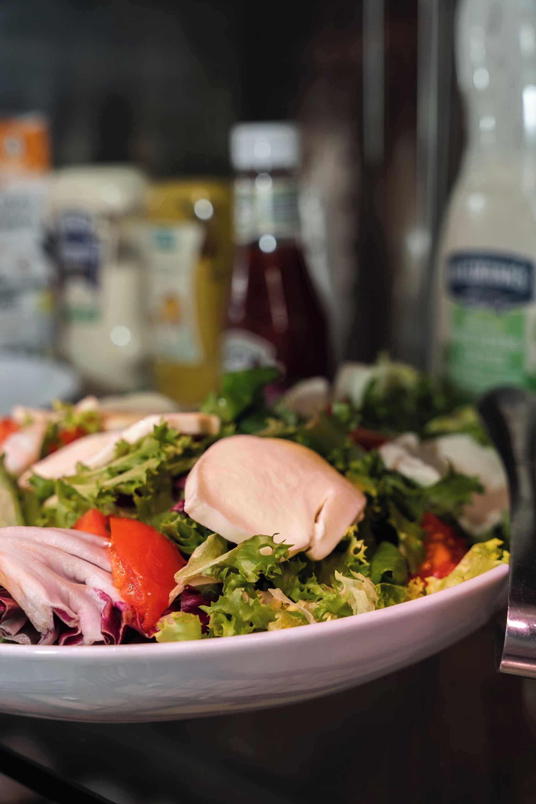 A bowl of salad with various vegetables and a piece of chicken, with condiments in the background.