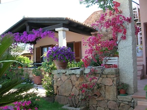 A stone wall with a potted plant and flowers, a wooden pergola, and a building with a tiled roof. - SVS - Isuledda Villas (San Teodoro)