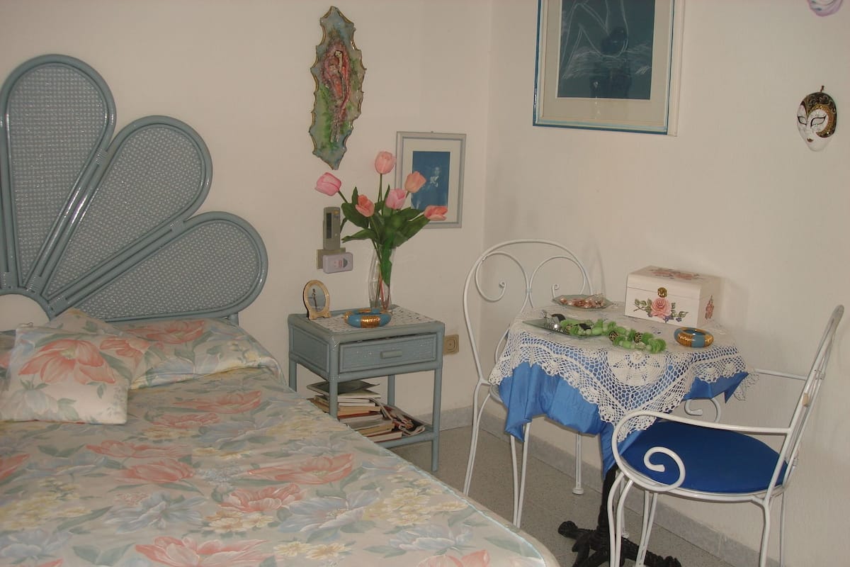 Un dormitorio con cama, mesita de noche, mesa con silla y pared con un cuadro y un espejo.