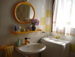 A bathroom with a white sink, yellow towel rack, and a mirror with a yellow frame. There are toiletries on the shelf, a flower vase, and a window with lace curtains. - SVS - Isuledda Villas (San Teodoro)
