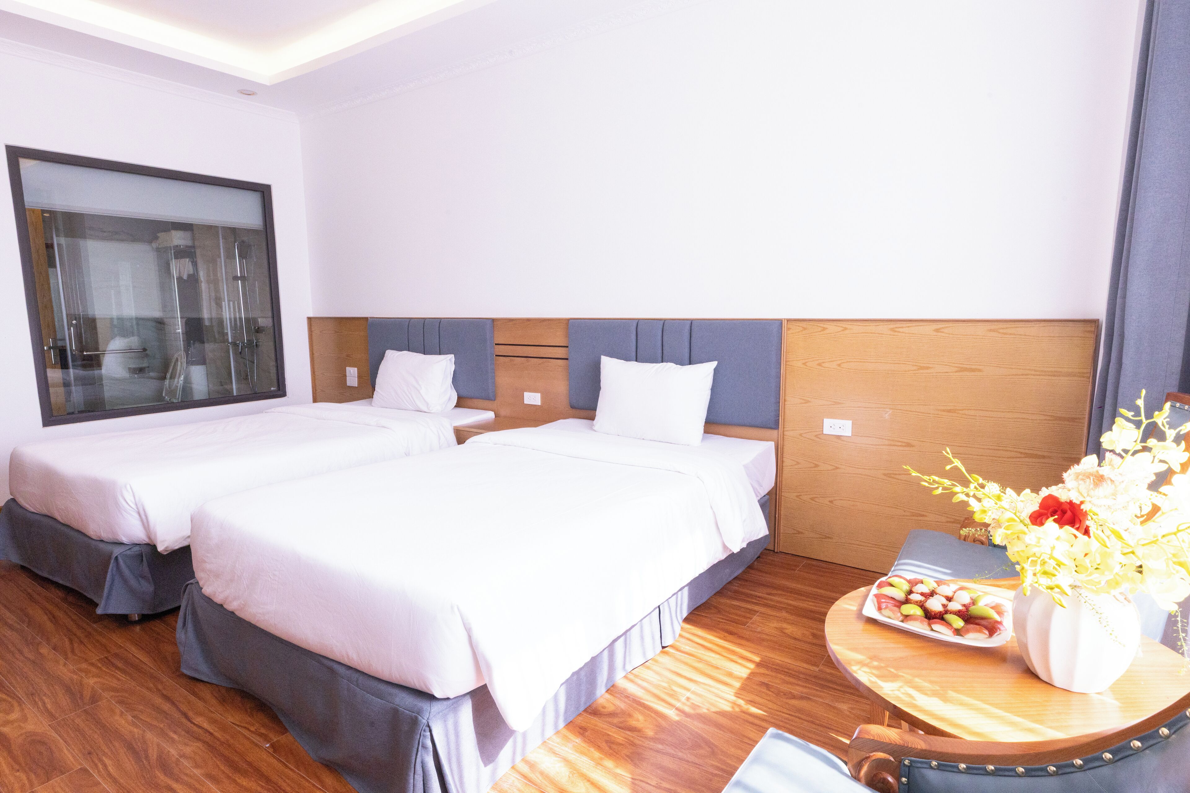 A hotel room with two beds, a wooden headboard, a small table with a vase of flowers, and a window with a view of the city.