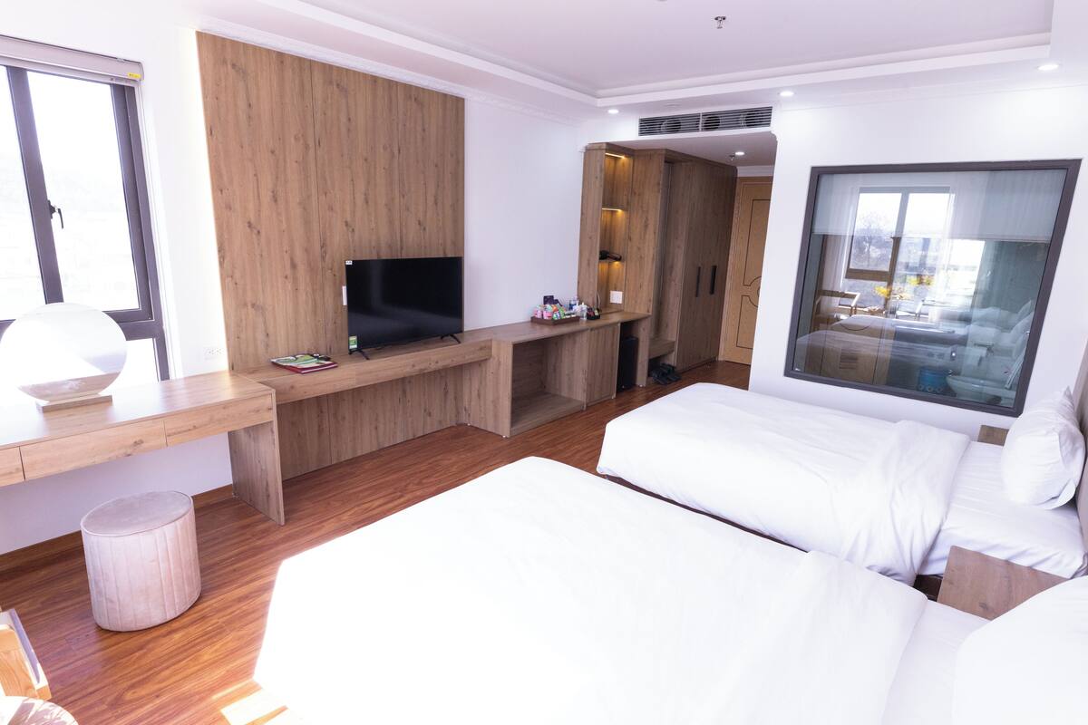 A modern hotel room with a wooden desk, a flat-screen TV, a bed with white linens, and a large window offering a view of the outdoors.