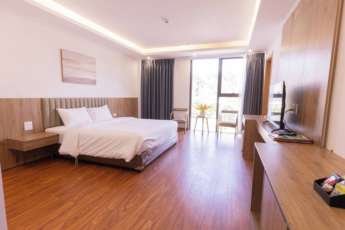 A modern hotel room with a wooden floor, a large bed with white linens, a desk with a flat-screen TV, and a balcony with a view.
