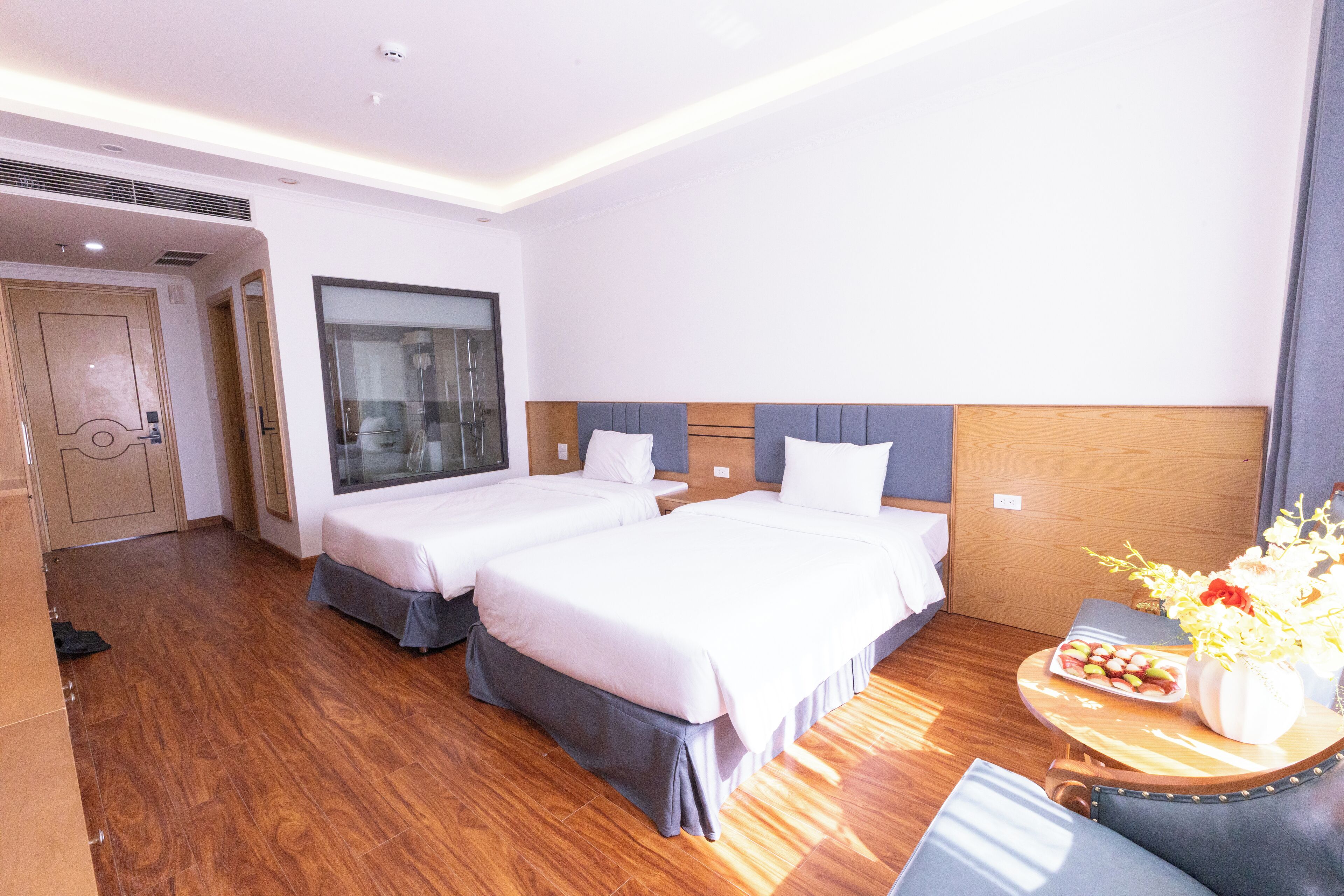 Habitación de hotel con dos camas, piso de madera, un ventanal grande y una mesita con un jarrón de flores.