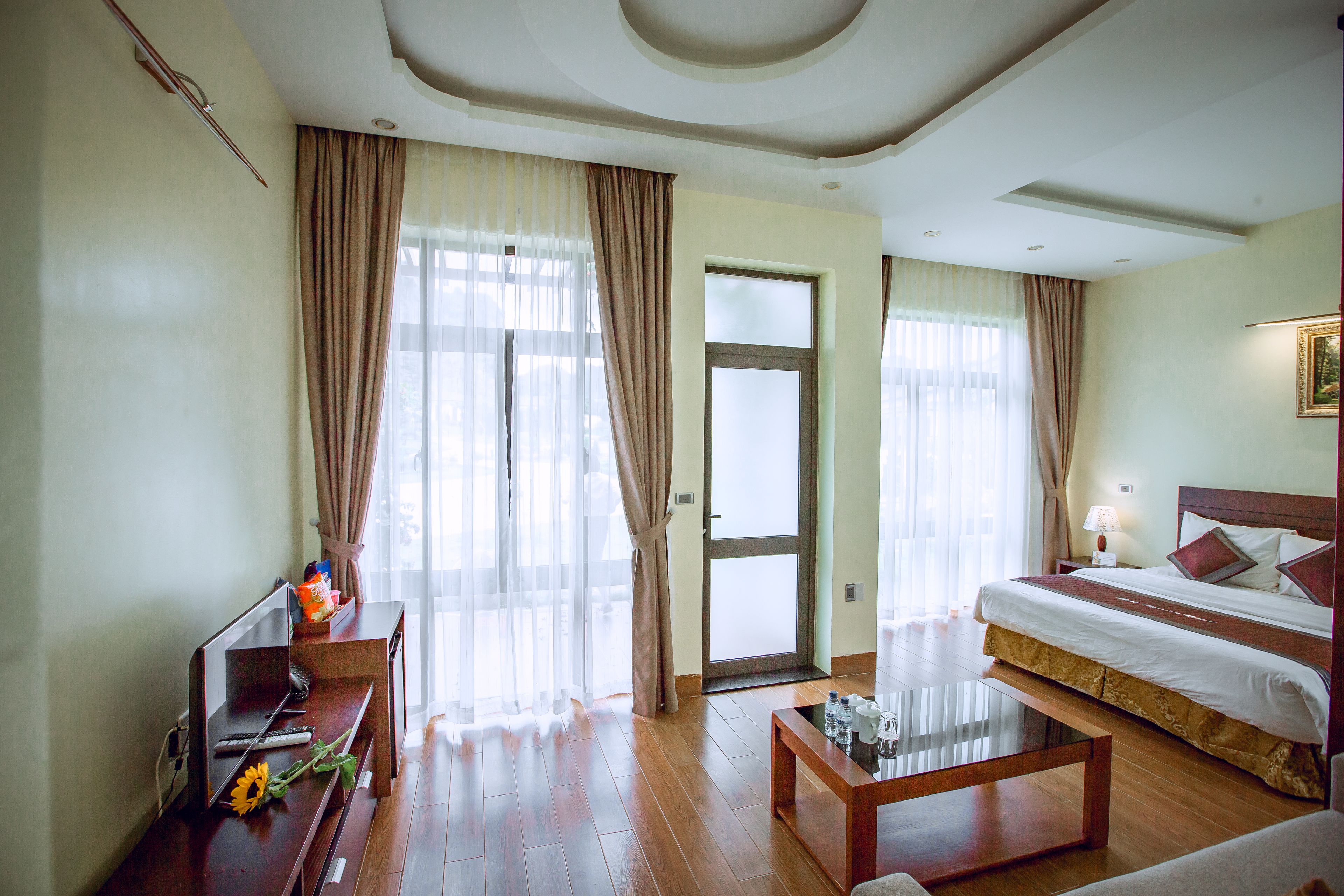 A hotel room with a large bed, a wooden coffee table, a desk with a computer and a sunflower, and a door leading to a balcony.