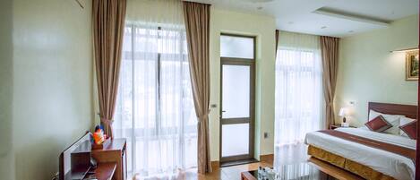 A hotel room with a large bed, a wooden coffee table, a desk with a computer and a sunflower, and a door leading to a balcony.