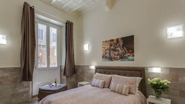 Un dormitorio con una cama grande, una mesita de noche, un jarrón con flores, un cuadro en la pared y una ventana con cortinas.