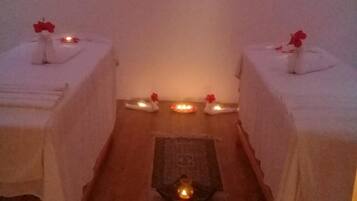 Two beds with white bedspreads and a red flower on each, accompanied by a lit candle and additional candles on the floor.