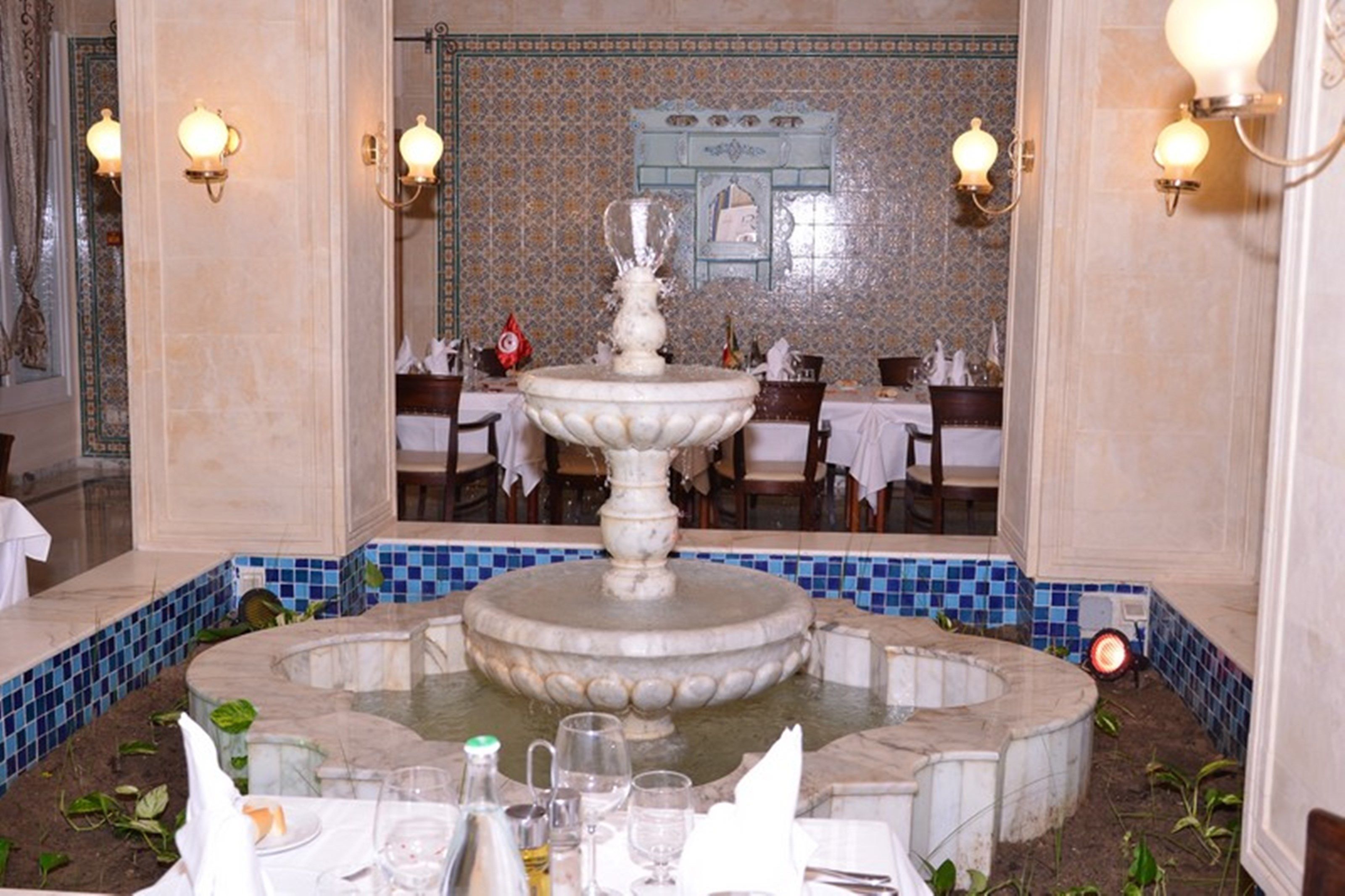 A dining area with a central fountain, tables set for dining, and a tiled wall with a decorative mirror.