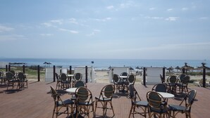 Un espace extérieur aménagé avec des chaises et des tables en osier, offrant une vue dégagée sur une plage, l’océan et un bateau au loin.