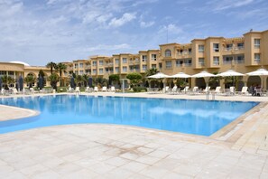 A hotel pool area with multiple lounge chairs and umbrellas, surrounded by palm trees and a building with balconies.