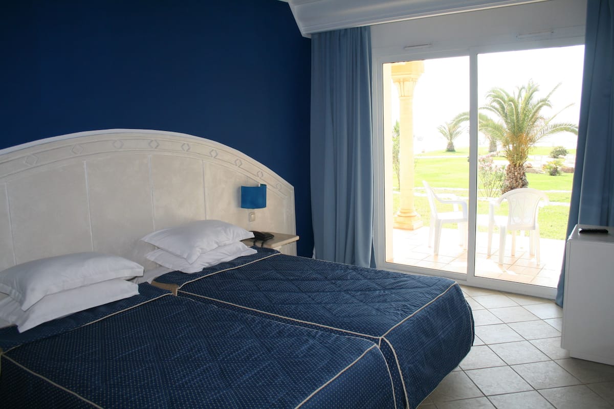 A bedroom with a large bed, a nightstand, and a view of a patio with chairs and a palm tree.