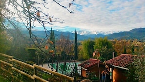 A mountainous landscape with a wooden fence, a small building with a red roof, and a body of water in the foreground. - Chambre d'hôtes St Jacques (Saint-Lizier)