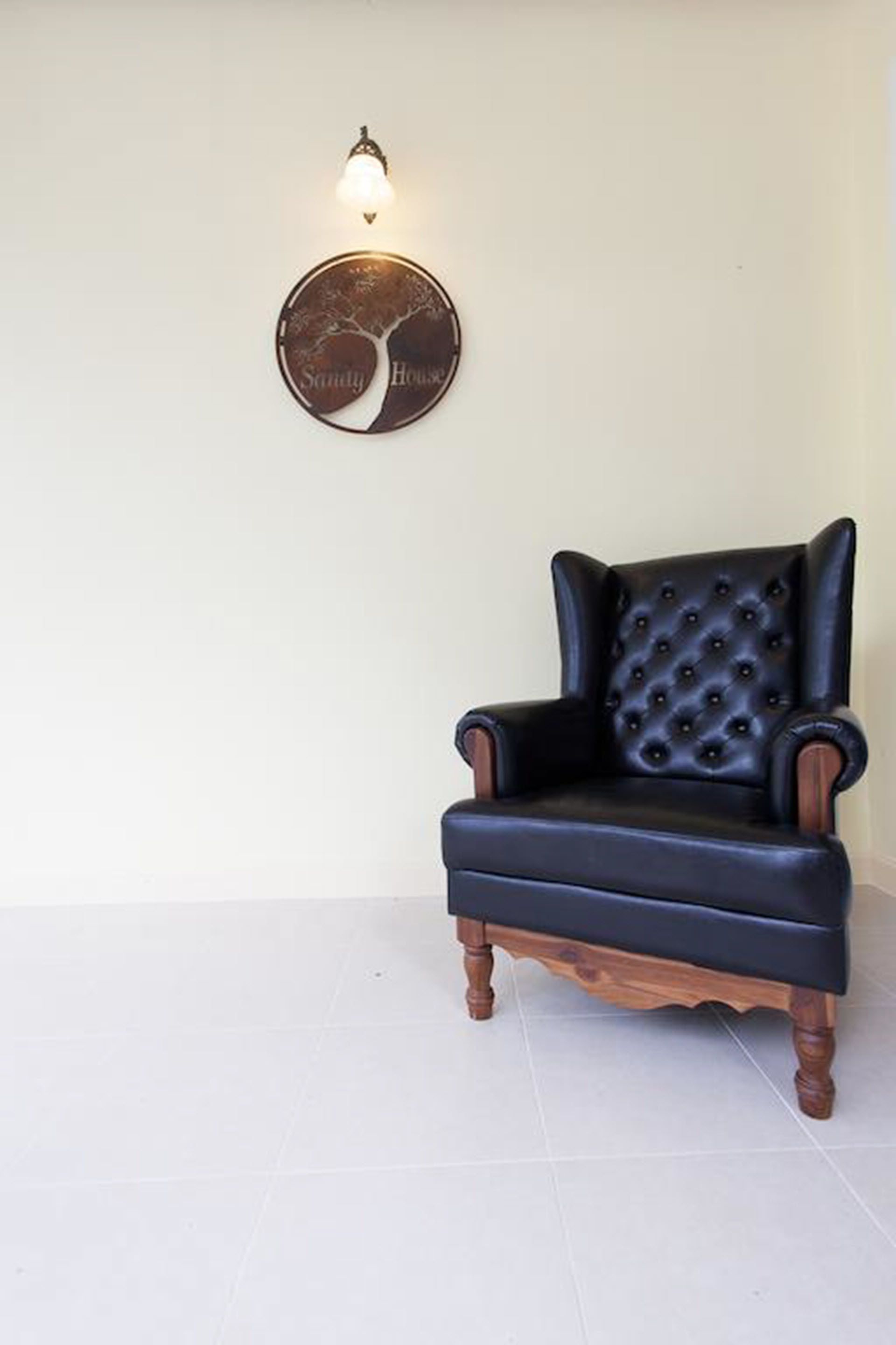 A black leather armchair with wooden legs and a tufted backrest, positioned in front of a wall-mounted round mirror with a tree design and the words "Sunny House" below it.