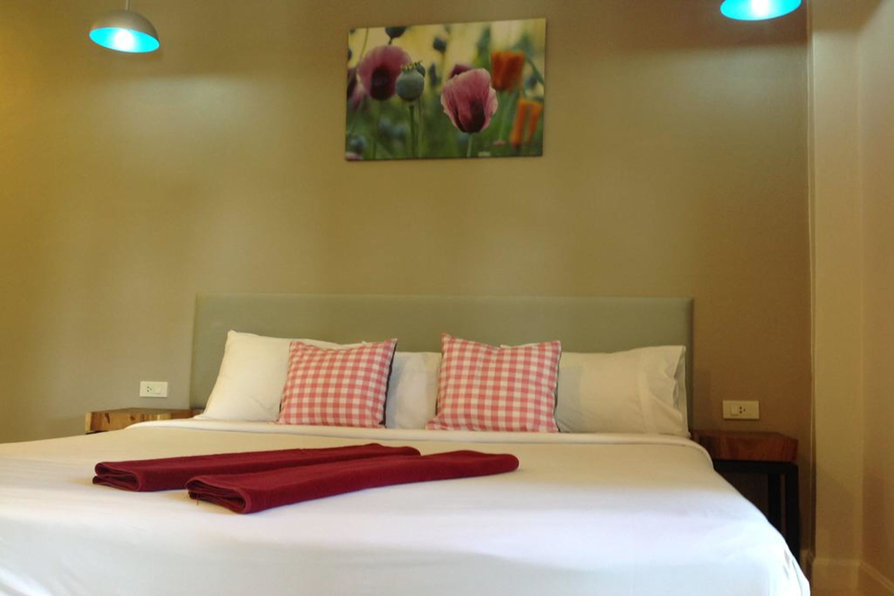 Un lit bien fait, avec du linge de lit blanc et des coussins à carreaux rouges, une serviette rouge posée sur le lit et un tableau de fleurs accroché au mur.