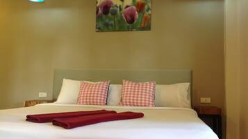 Un lit bien fait, avec du linge de lit blanc et des coussins à carreaux rouges, une serviette rouge posée sur le lit et un tableau de fleurs accroché au mur.